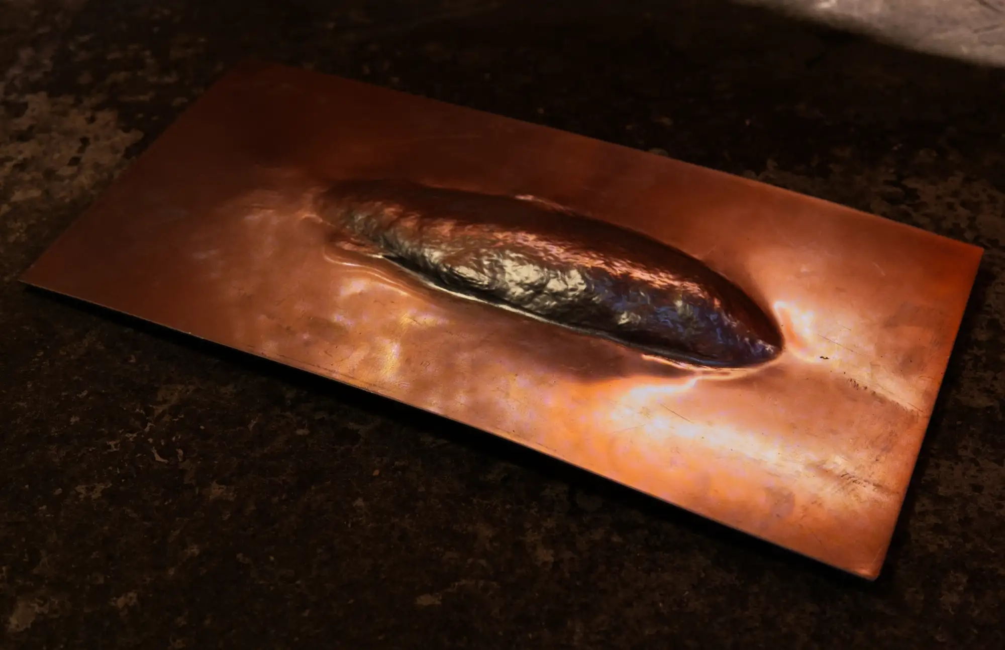 Ein Kupferblech, in dem sich ein fischförmiges Objekt abdrückt.
