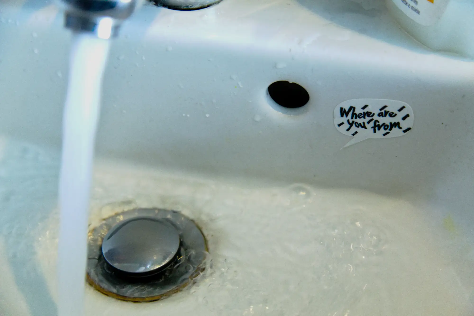 Ein Waschbecken, aus dem Wasserhahn fließt Wasser, im Abfluss verschwindet es. daneben ein Sticker: Where are you from?