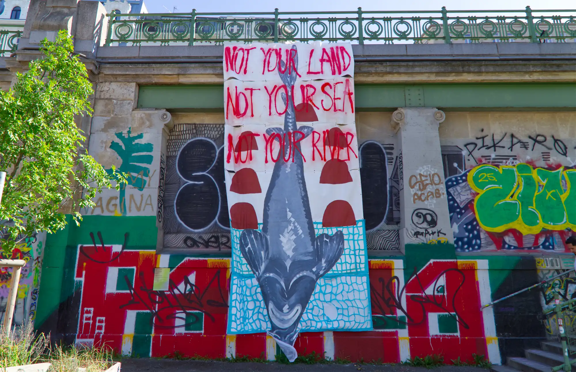 Ein Banner hängt von der Promenade am Donaukanal. Darauf ein Grinsender Fisch und die Wirte: NOT YOUR LAND, NOT YOUR SEA, NOT YOUR RIVER.