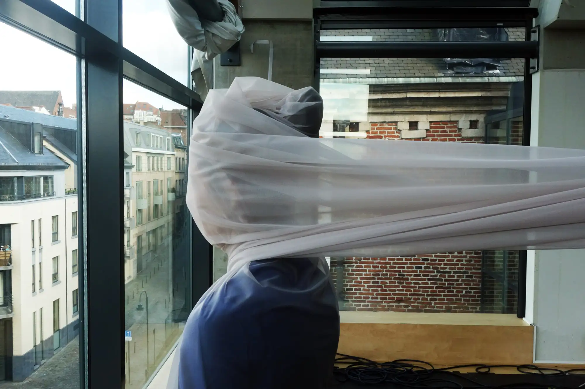 Person sieht aus einem Fenster und hat ein transparentes Tuch um den Kopf gewickelt