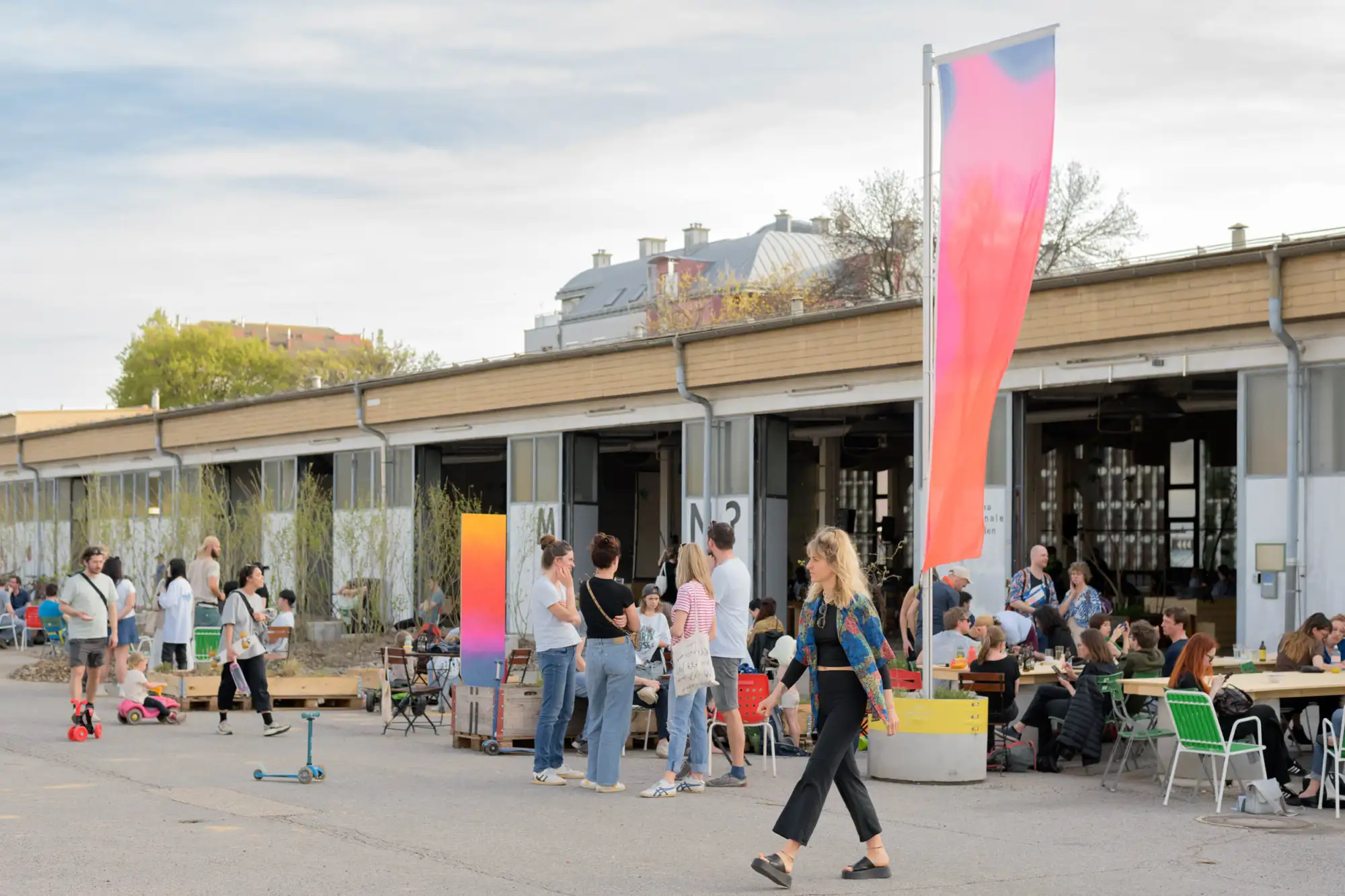 Eine lebhafte Szene im Freien vor einem Industriegebäude: Menschen sitzen an langen Holztischen, essen, trinken und unterhalten sich. Andere stehen in kleinen Gruppen oder gehen vorbei. Bunte Fahnen und Schilder mit Farbverläufen schmücken den Platz, während Kinder mit kleinen Fahrzeugen spielen.